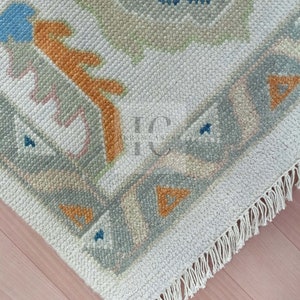 May include: A close-up of a white rug with a geometric pattern in shades of blue, orange, and grey. The rug has a fringe border.