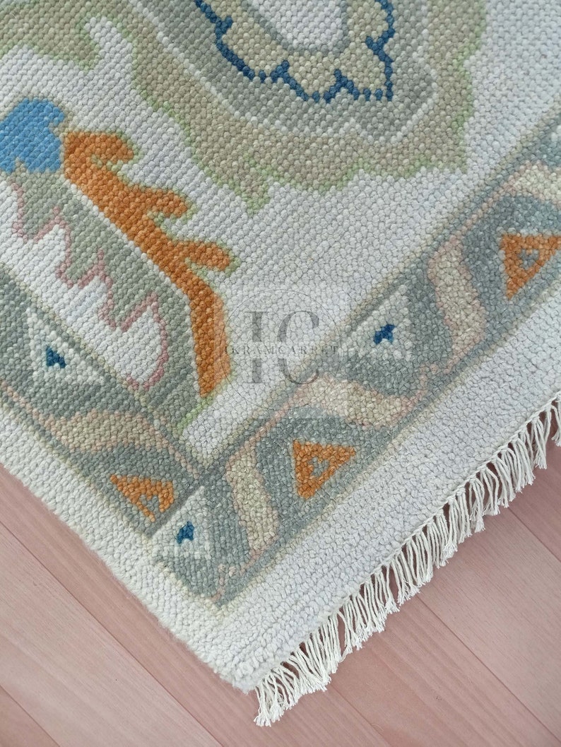 May include: A close-up of a white rug with a geometric pattern in shades of blue, orange, and grey. The rug has a fringe border.