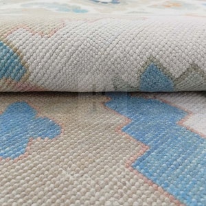 May include: Close-up of a beige and white rug with a geometric pattern in shades of blue and pink. The rug has a soft, textured surface.