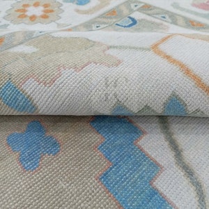 May include: Close-up of a white rug with a geometric pattern in shades of blue, orange, and pink. The rug has a soft, textured surface.