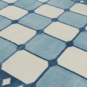 May include: A flat-woven rug featuring a repeating grid pattern of alternating light blue and off-white squares. The intersections of the grid are marked by small, dark blue geometric star-like motifs that connect the larger square sections.