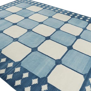 May include: A rectangular area rug featuring a repeating grid pattern of alternating light blue and off-white squares. The design includes a dark blue border decorated with a series of small, white, diamond-shaped geometric motifs along the edges.
