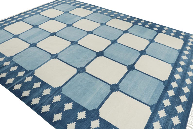 May include: A rectangular area rug featuring a repeating grid pattern of alternating light blue and off-white squares. The design includes a dark blue border decorated with a series of small, white, diamond-shaped geometric motifs along the edges.