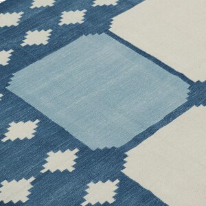 May include: A woven area rug featuring a geometric pattern of large squares in shades of dark blue, light blue, and off-white. The design includes small diamond-shaped motifs along the dark blue border and white fringe at the corner edge.