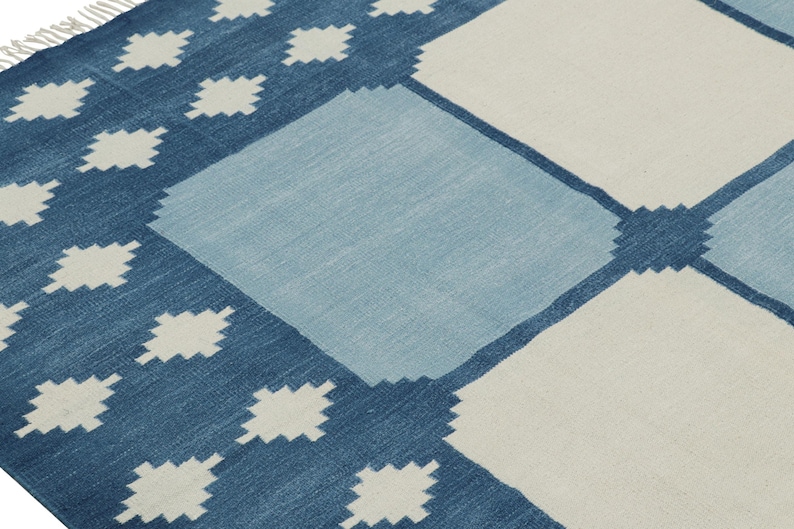 May include: A woven area rug featuring a geometric pattern of large squares in shades of dark blue, light blue, and off-white. The design includes small diamond-shaped motifs along the dark blue border and white fringe at the corner edge.