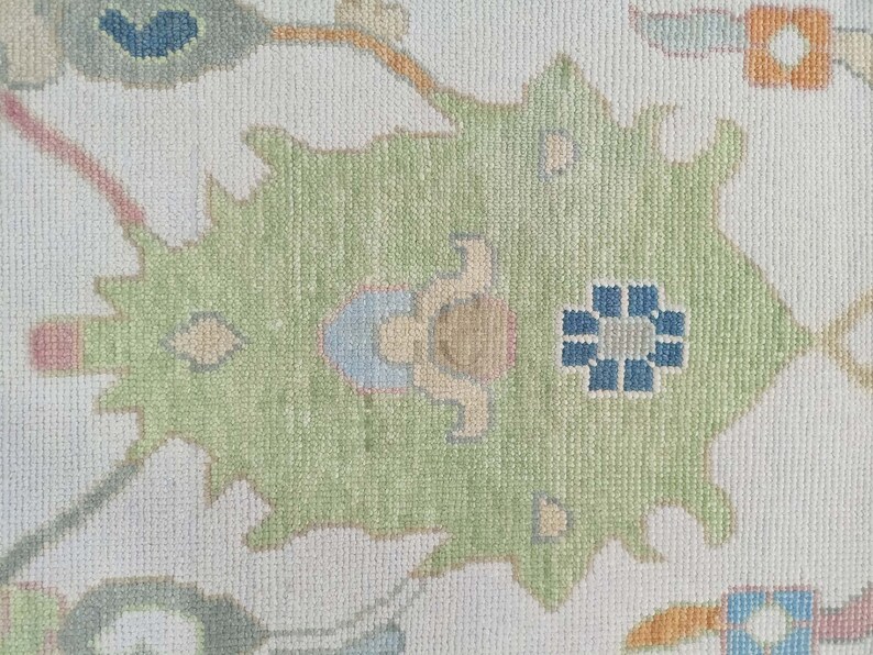 May include: A close-up of a cream-coloured rug with a green, blue, and orange floral pattern. The rug has a subtle texture and a traditional design.