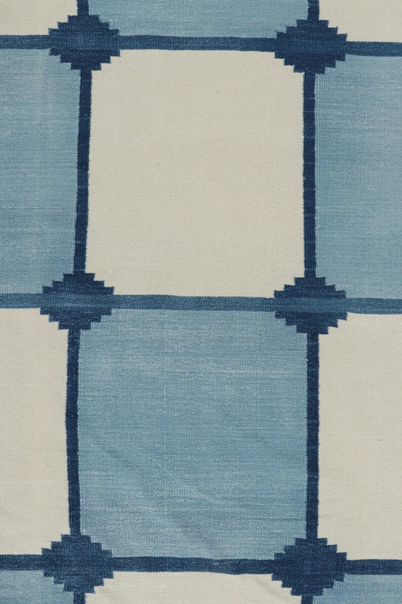 May include: A woven textile featuring a grid pattern of alternating light blue and off-white squares. The grid lines are defined by dark navy blue horizontal and vertical bands, with small, stepped, cross-shaped motifs at each intersection.