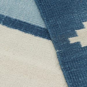 May include: A woven textile rug featuring a geometric pattern in shades of dark blue, light blue, and off-white. The fabric displays a visible textured weave with a stepped, jagged design element on the right side and horizontal color blocking.