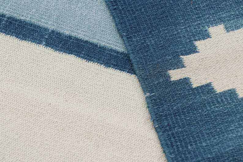 May include: A woven textile rug featuring a geometric pattern in shades of dark blue, light blue, and off-white. The fabric displays a visible textured weave with a stepped, jagged design element on the right side and horizontal color blocking.