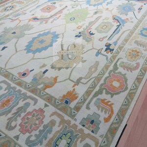 May include: A cream-coloured rug with a traditional Persian design. The rug features intricate patterns in shades of blue, green, pink, and orange. The rug has a fringed border.