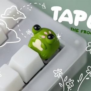 Puede incluir: Una tecla con forma de rana verde con ojos negros y mejillas rosas está instalada en un teclado blanco. La tecla está etiquetada como "TAPU THE FROG" en texto blanco y verde. Garabatos caprichosos de nubes y flores decoran el fondo.