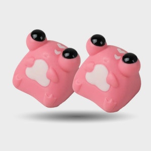May include: Two pink frog-shaped objects with large, black eyes and white bellies. The objects have a smooth texture and are slightly angled, casting a soft shadow. The design is cartoonish and playful.
