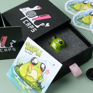 May include: A green frog-shaped keycap, "TAPU the Frog", is presented in a black box with the "LOL CAPS" logo. The box is open, revealing the keycap on a foam insert. Stickers and pins with frog illustrations are also visible.