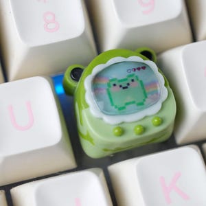 K&ouml;nnte beinhalten: Ein gr&uuml;ner, froschf&ouml;rmiger Keycap mit einem pixeligen Froschdesign auf einer wei&szlig;en und rosa Tastatur. Der Keycap hat einen wei&szlig;en, gewellten Rand und drei gr&uuml;ne Kn&ouml;pfe. Die Tastaturtasten sind wei&szlig; mit rosa Buchstaben.