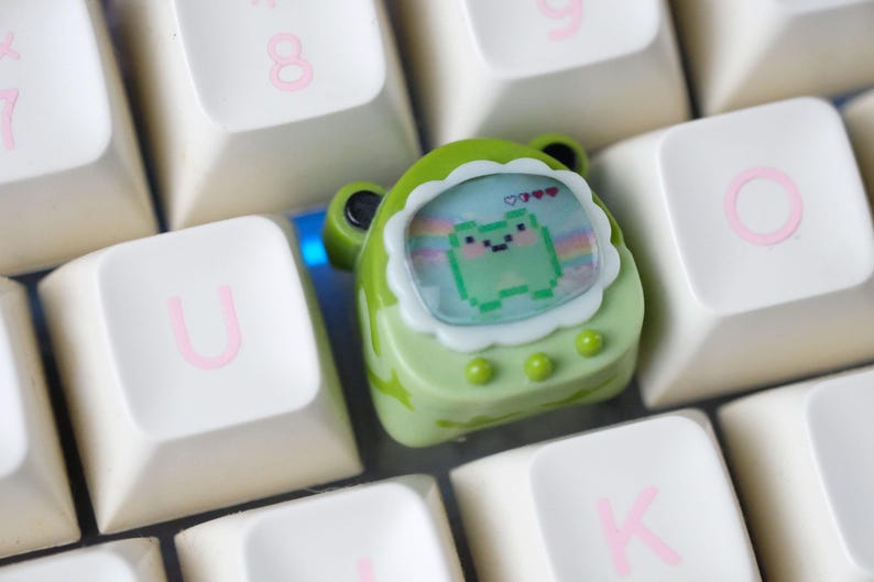 K&ouml;nnte beinhalten: Ein gr&uuml;ner, froschf&ouml;rmiger Keycap mit einem pixeligen Froschdesign auf einer wei&szlig;en und rosa Tastatur. Der Keycap hat einen wei&szlig;en, gewellten Rand und drei gr&uuml;ne Kn&ouml;pfe. Die Tastaturtasten sind wei&szlig; mit rosa Buchstaben.