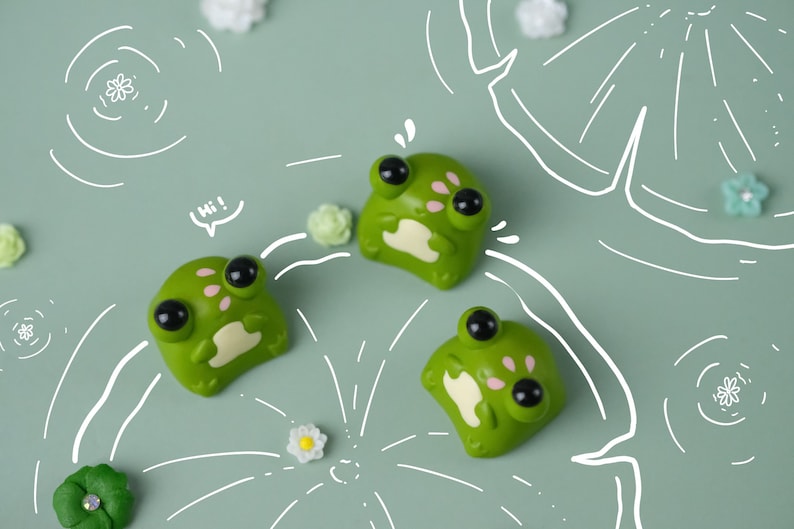 May include: Three green frog figurines with large black eyes and pink accents are arranged on a light green surface. The word "Hi!" is in a speech bubble. White line drawings of lily pads and flowers are also present.