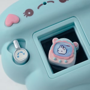 K&ouml;nnte beinhalten: Ein hellblaues elektronisches Spielzeug mit einem Bildschirm, der einen verpixelten Hasencharakter anzeigt. Das Spielzeug hat rosa und braune Akzente, eine herzf&ouml;rmige Taste und ein silbernes Zifferblatt. Der Bildschirm zeigt einen Hasencharakter.