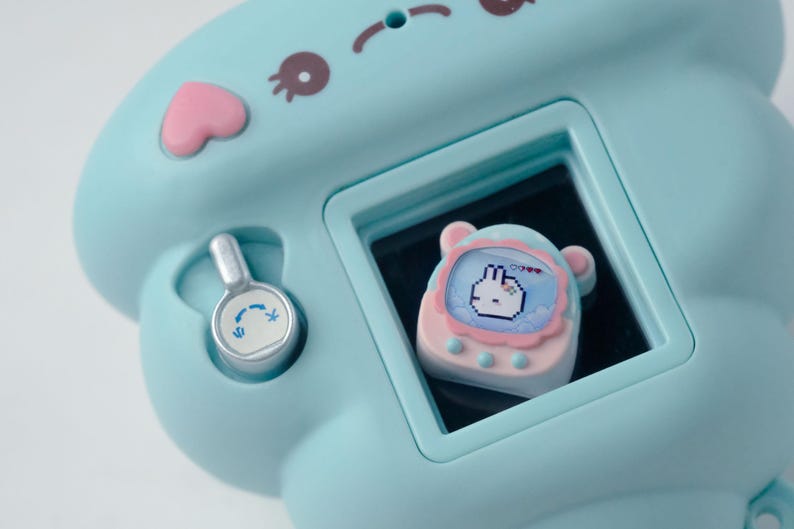 K&ouml;nnte beinhalten: Ein hellblaues elektronisches Spielzeug mit einem Bildschirm, der einen verpixelten Hasencharakter anzeigt. Das Spielzeug hat rosa und braune Akzente, eine herzf&ouml;rmige Taste und ein silbernes Zifferblatt. Der Bildschirm zeigt einen Hasencharakter.