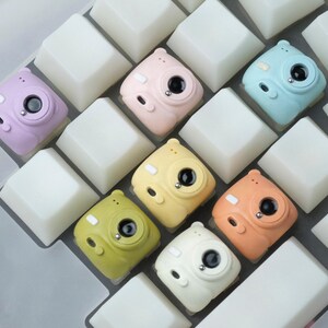 Peut inclure: Un ensemble de touches de clavier miniatures en forme d'appareil photo, aux couleurs pastel lavande, rose, bleu, jaune, vert, orange et blanc. Les touches sont disposées parmi des touches de clavier blanches, créant une esthétique ludique et unique.