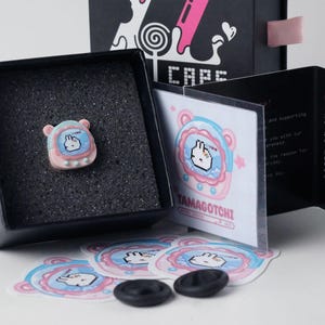 K&ouml;nnte beinhalten: Ein Tamagotchi-Pin in Pastellfarben wird in einer schwarzen Box mit rosa Band pr&auml;sentiert. Aufkleber und ein Anleitungsheft sind ebenfalls sichtbar. Die Box ist mit "CAPS" beschriftet.