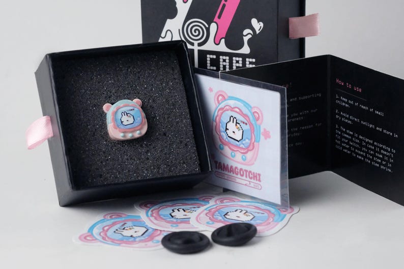 K&ouml;nnte beinhalten: Ein Tamagotchi-Pin in Pastellfarben wird in einer schwarzen Box mit rosa Band pr&auml;sentiert. Aufkleber und ein Anleitungsheft sind ebenfalls sichtbar. Die Box ist mit "CAPS" beschriftet.
