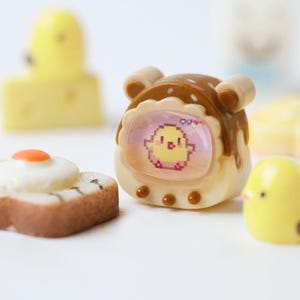 K&ouml;nnte beinhalten: Eine Sammlung von Miniatur-Lebensmittel- und Charakterfiguren. Enth&auml;lt einen braunen und wei&szlig;en Toast mit Spiegelei, ein gelbes K&uuml;ken, einen gelben K&auml;seblock und ein braunes und wei&szlig;es Ger&auml;t mit einem pixeligen K&uuml;ken auf dem Bildschirm.