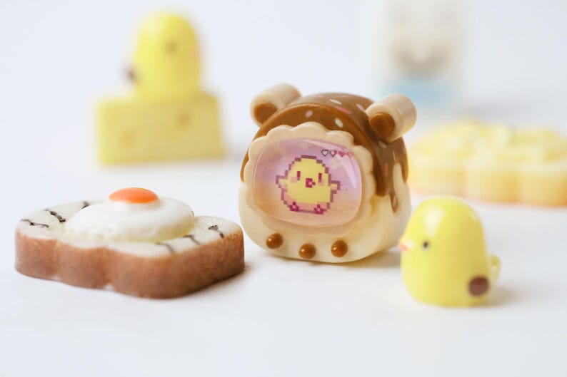 K&ouml;nnte beinhalten: Eine Sammlung von Miniatur-Lebensmittel- und Charakterfiguren. Enth&auml;lt einen braunen und wei&szlig;en Toast mit Spiegelei, ein gelbes K&uuml;ken, einen gelben K&auml;seblock und ein braunes und wei&szlig;es Ger&auml;t mit einem pixeligen K&uuml;ken auf dem Bildschirm.