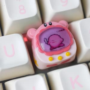 K&ouml;nnte beinhalten: Eine rosa-wei&szlig;e, individuelle Tastenkappe mit einer Cartoonfigur auf einem Bildschirm, umrahmt von Rosa und gelben Akzenten. Die Tastenkappe befindet sich auf einer wei&szlig;en Tastatur mit rosa Buchstaben.