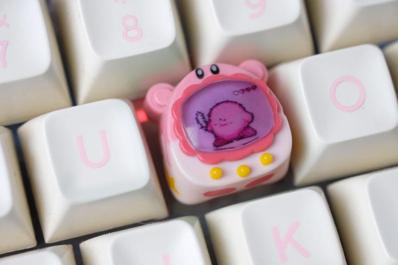 K&ouml;nnte beinhalten: Eine rosa-wei&szlig;e, individuelle Tastenkappe mit einer Cartoonfigur auf einem Bildschirm, umrahmt von Rosa und gelben Akzenten. Die Tastenkappe befindet sich auf einer wei&szlig;en Tastatur mit rosa Buchstaben.