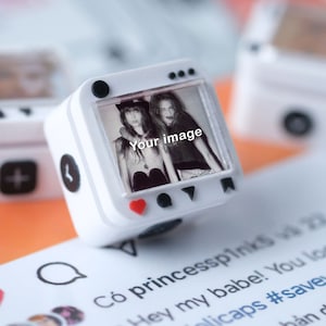 May include: White, square ring with a small screen showing a black and white photo of two women. The ring features a heart-shaped button and Instagram-style icons. The text "Your image" is visible on the screen.