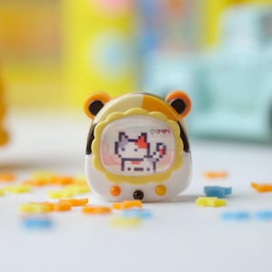 K&ouml;nnte beinhalten: Ein tragbares elektronisches Haustierspielzeug mit einem pixeligen Katzencharakter auf dem Bildschirm. Das Spielzeug ist wei&szlig;, gelb und braun, mit orangefarbenen, ohrenartigen Akzenten. Bunte stern- und bl&uuml;tenf&ouml;rmige Dekorationen sind verstreut.