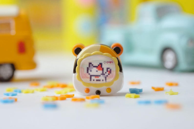 K&ouml;nnte beinhalten: Ein tragbares elektronisches Haustierspielzeug mit einem pixeligen Katzencharakter auf dem Bildschirm. Das Spielzeug ist wei&szlig;, gelb und braun, mit orangefarbenen, ohrenartigen Akzenten. Bunte stern- und bl&uuml;tenf&ouml;rmige Dekorationen sind verstreut.