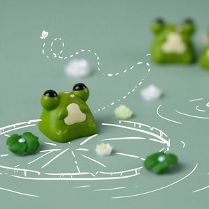 May include: A charming image of a green frog figurine with black eyes, placed on a lily pad. The background is a soft green, with other frog figurines and decorative elements. The scene has a playful, nature-inspired theme, reminiscent of a pond.