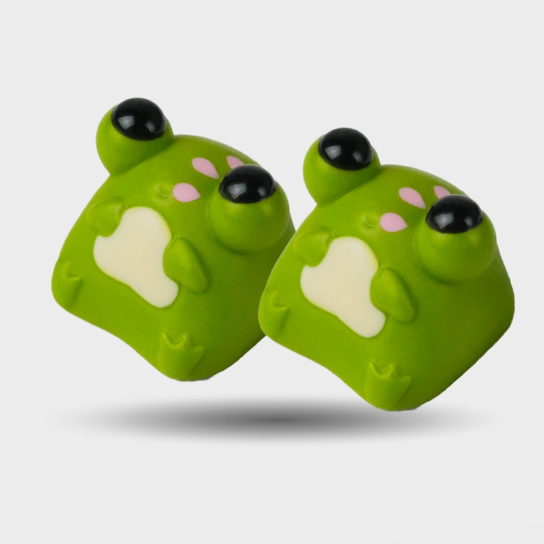 May include: Two green frog-shaped decorative items with black eyes and pink accents. The frog-shaped items have a white patch on their chest. The items are designed to look like cartoon frogs.
