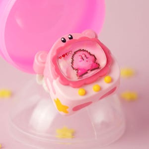 K&ouml;nnte beinhalten: Ein rosa-wei&szlig;es Spielzeug mit einem Kirby-Charakterdesign. Das Spielzeug befindet sich in einem transparenten Kunststoffbeh&auml;lter mit gelben sternf&ouml;rmigen Dekorationen. Das Spielzeug hat einen Bildschirm, der einen pixeligen Kirby-Charakter anzeigt.