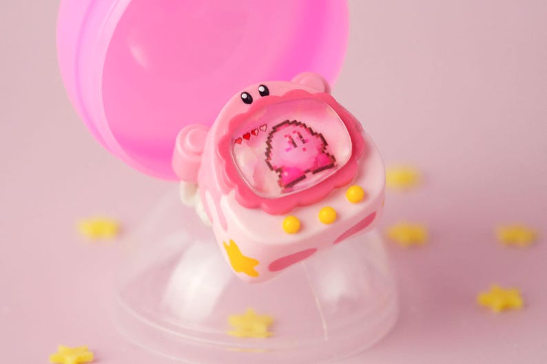 K&ouml;nnte beinhalten: Ein rosa-wei&szlig;es Spielzeug mit einem Kirby-Charakterdesign. Das Spielzeug befindet sich in einem transparenten Kunststoffbeh&auml;lter mit gelben sternf&ouml;rmigen Dekorationen. Das Spielzeug hat einen Bildschirm, der einen pixeligen Kirby-Charakter anzeigt.