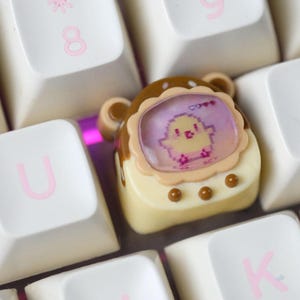 K&ouml;nnte beinhalten: Eine individuelle Tastenkappe mit einem pixeligen K&uuml;ken-Design in einem dekorativen Rahmen. Die Kappe ist cremefarben mit braunen Akzenten und befindet sich zwischen wei&szlig;en Tastaturtasten mit rosa Buchstaben.
