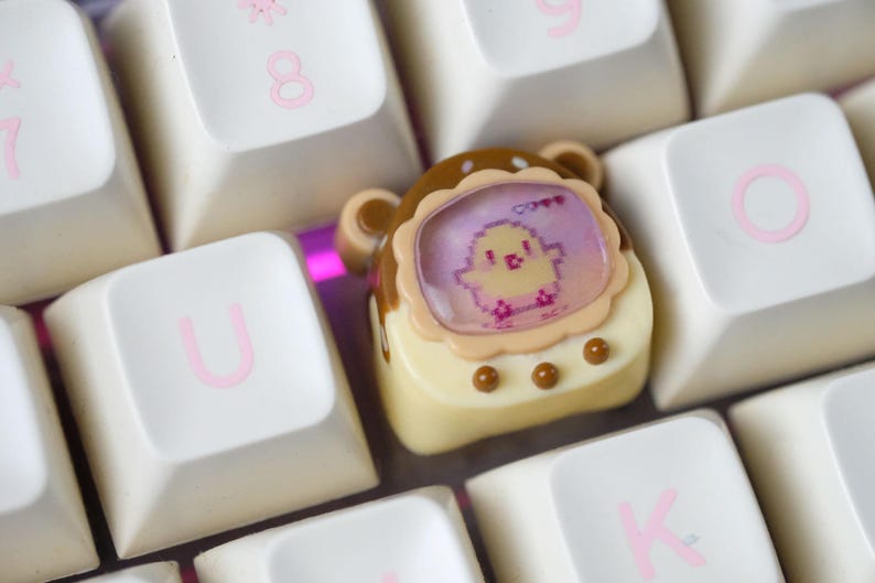 K&ouml;nnte beinhalten: Eine individuelle Tastenkappe mit einem pixeligen K&uuml;ken-Design in einem dekorativen Rahmen. Die Kappe ist cremefarben mit braunen Akzenten und befindet sich zwischen wei&szlig;en Tastaturtasten mit rosa Buchstaben.