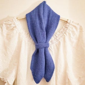 May include: A blue knitted scarf tied in a bow, hanging on a white hanger against a white wall.