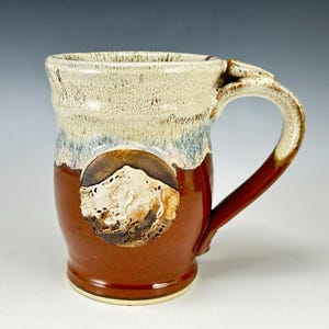May include: Handmade ceramic mug with a mountain design. The mug features a brown base, a cream-colored rim, and a dark red handle. The mountain design is in a circular shape on the front of the mug.