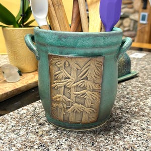 May include: A teal ceramic utensil holder with a bamboo design. The holder has two handles and is filled with wooden spoons and purple and white spatulas. The bamboo design is a light brown color.
