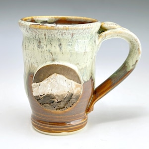 May include: A handmade ceramic mug with a mountain design. The mug features a light green and brown glaze, with a circular mountain relief. The handle and rim have a unique shape. The mug is approximately 4 inches tall.
