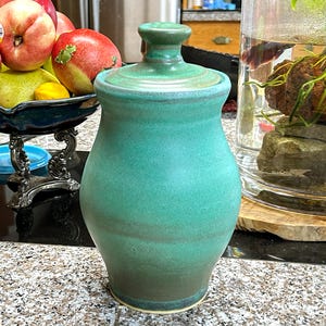 May include: A teal ceramic jar with a lid. The jar has a rounded body and a slightly flared base. The lid is a matching teal color and has a small knob on top. The jar is sitting on a speckled countertop.