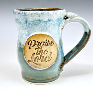 May include: A ceramic mug with a blue and white drip glaze finish. The mug features a circular tan medallion on the front with the embossed text Praise the Lord in a script font. It has a curved handle and a flared rim with a smooth texture.