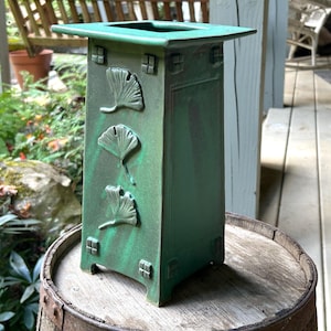 May include: A square, emerald green ceramic vase with a flared top rim. The front panel features three raised ginkgo leaf motifs arranged vertically, accented by small square decorative tiles at the top and bottom corners of the vase structure.