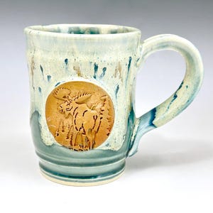 May include: Handmade ceramic mug with a light blue and cream glaze. The mug features a circular, raised relief of a moose in a forest scene. The handle and base have a darker blue-green color, adding contrast to the design.