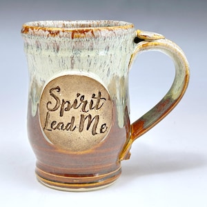 May include: A ceramic mug with a glossy finish featuring a two-tone glaze of cream and brown. The upper portion has a cream drip effect over a brown base. A circular medallion on the front is embossed with the text Spirit Lead Me in script.