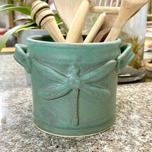 May include: A cylindrical ceramic utensil holder in a matte sage green glaze featuring a raised dragonfly relief on the front and two small side handles. The container is filled with several light-colored wooden kitchen tools.