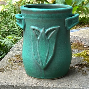 May include: A teal ceramic vessel with a wide circular opening, two side handles, and a raised relief design of a stylized tulip flower on the front. The surface features a matte finish with subtle variations in the deep green and blue tones.