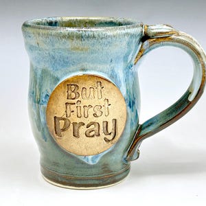 May include: A handmade ceramic mug with a blue and green glaze. The mug features a circular, tan-colored emblem with the words "But First Pray" embossed on the front. The handle is a darker shade of blue and brown.
